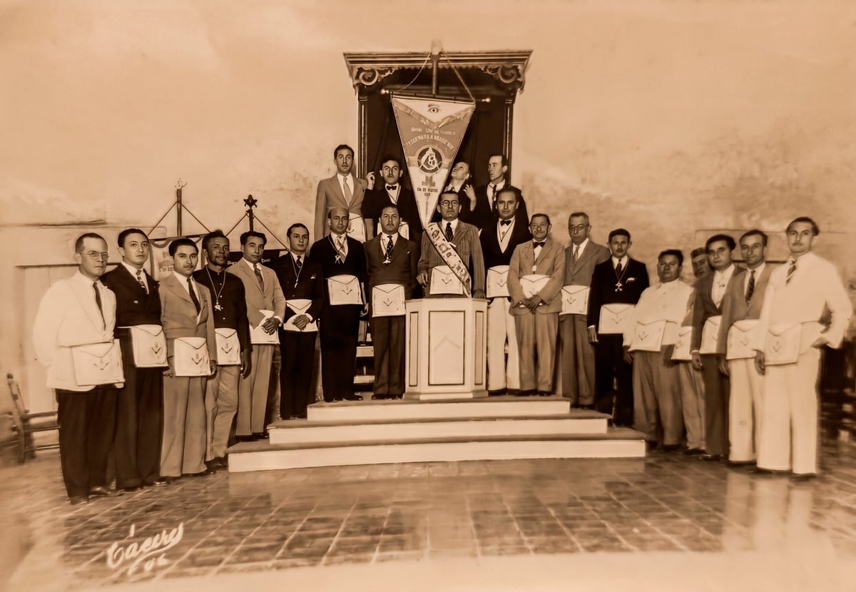 Sepia-toned vintage photograph of a large group of men in formal attire posed on and around a ceremonial platform with a banner.