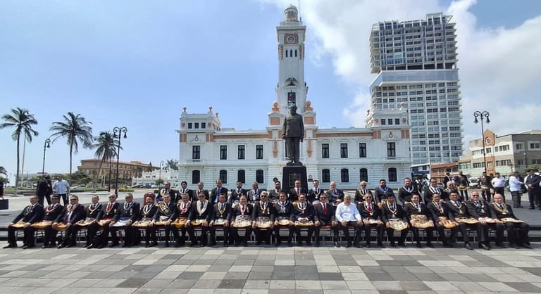 Large group of people in formal attire seated in rows facing a historic colonial building with tower and palm trees in a plaza