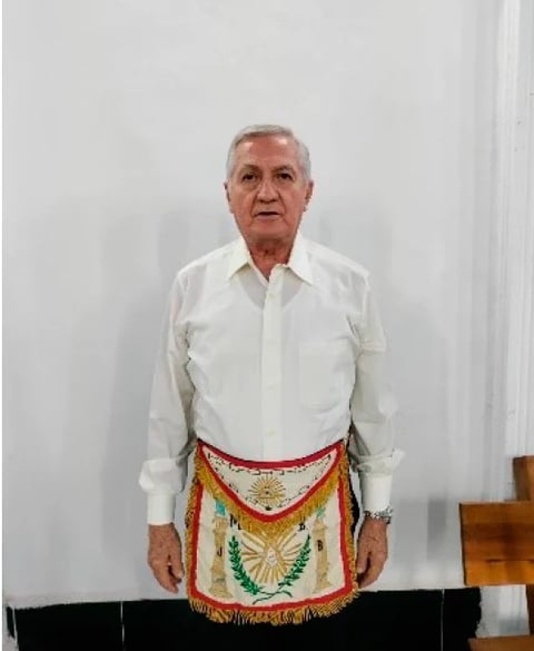 Older man wearing white dress shirt and colorful embroidered ceremonial apron standing against white wall