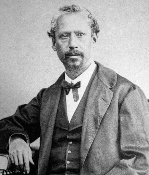 Black and white portrait photograph of a bearded man in formal Victorian-era attire with bow tie and three-piece suit, seated and resting his arm