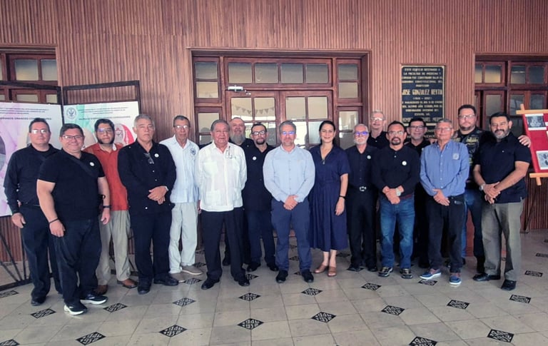 Grupo de aproximadamente 20 personas frente al edificio de ladrillo con láminas rojas de la Facultad de Medicina de la UADY, inaugurando la exposición Masones forjadores