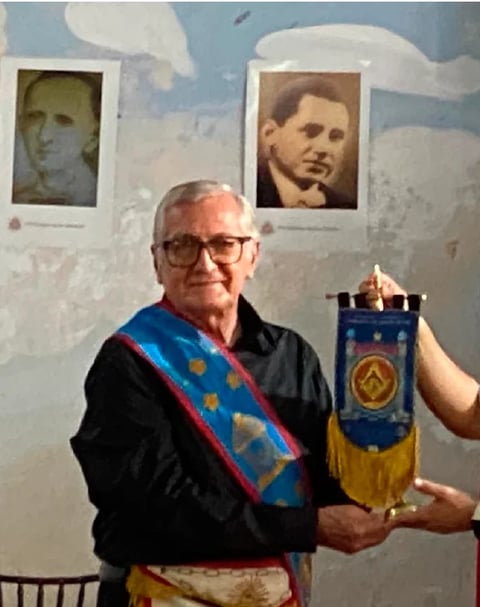 Elderly man wearing glasses and a blue and yellow sash holding a ceremonial banner with a star emblem, standing in front of two historical portrait photographs