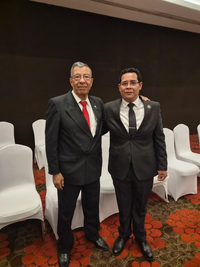 Two men in formal business suits posing together at an indoor event with white covered chairs and red patterned carpet visible in the background