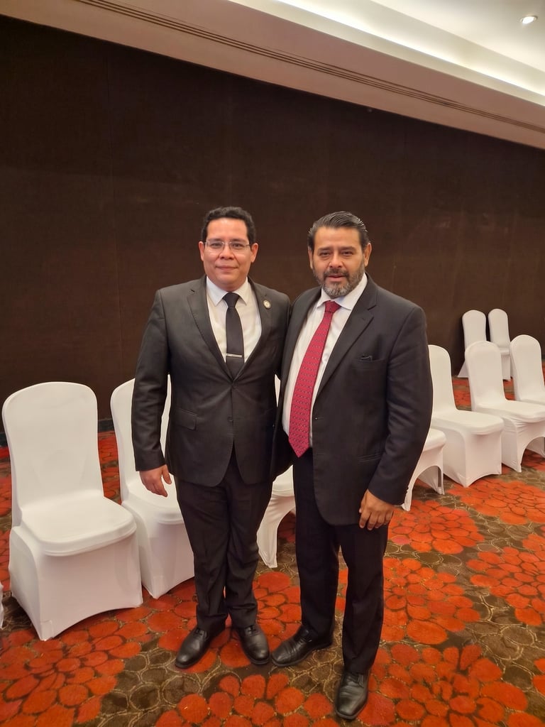 Two men in business suits standing together in an elegant venue with white chairs and orange patterned carpet in the background