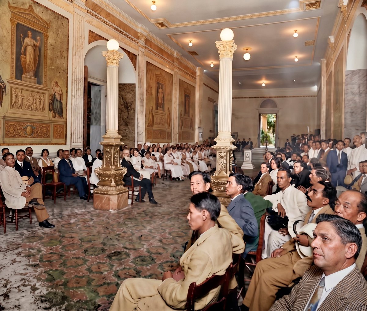 Large gathering of formally dressed attendees seated in an ornate hall with classical columns and gold-trimmed walls.